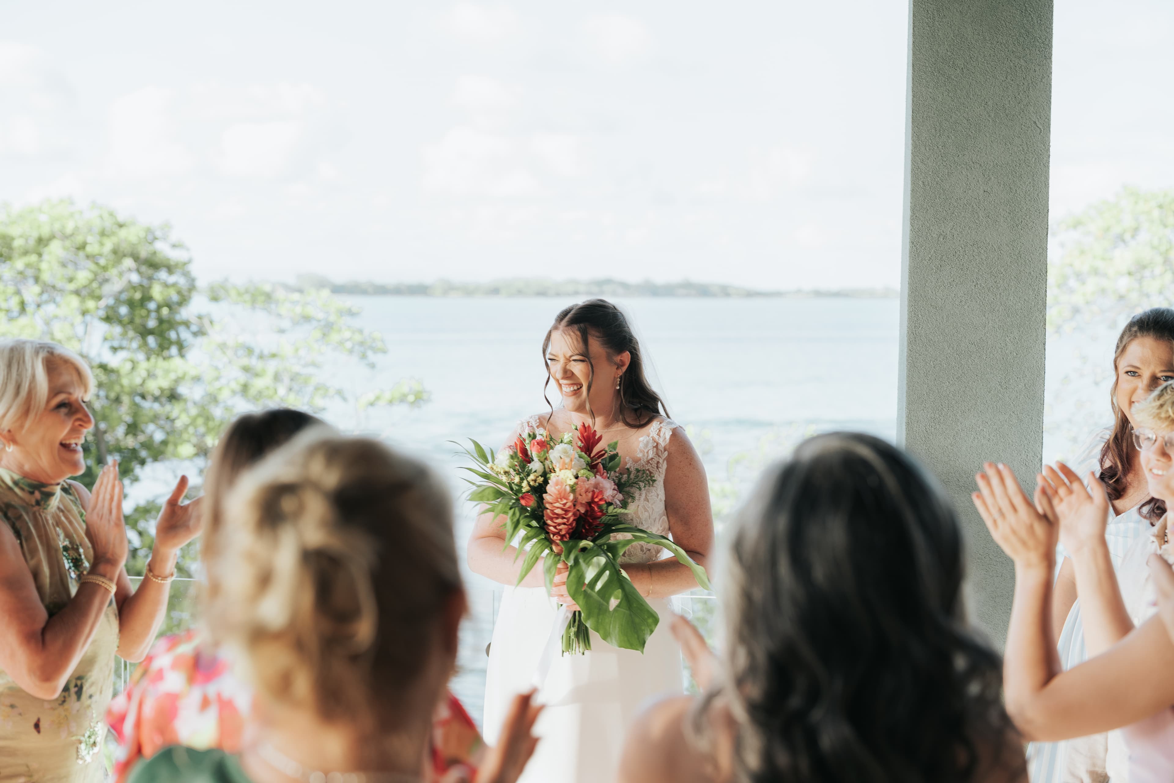 A smiling bride holds a vibrant bouquet while surrounded by applauding friends against a scenic waterfront backdrop.