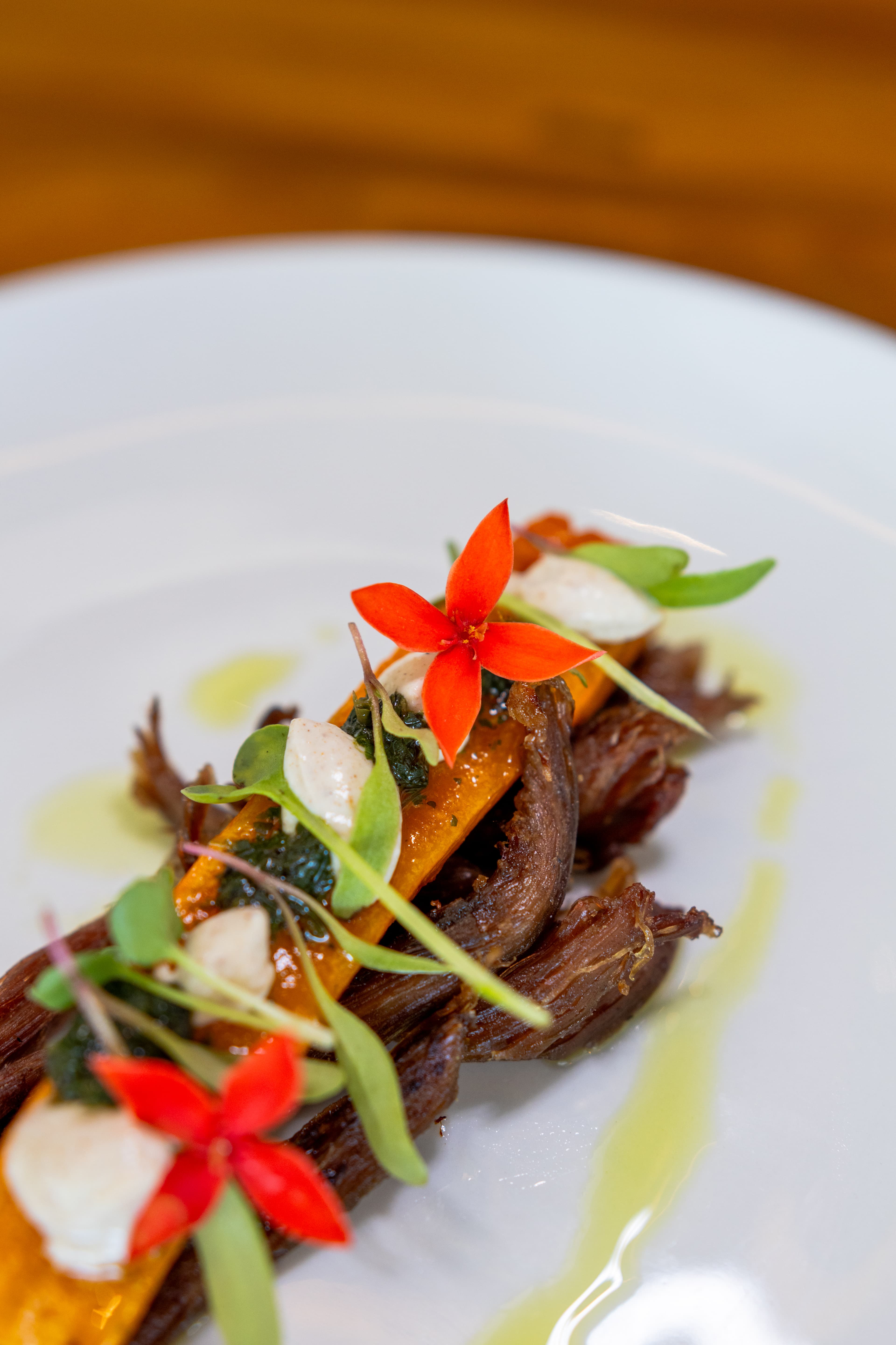 A beautifully plated dish featuring pulled beef, orange squash, creamy dollops, and vibrant red flowers.
