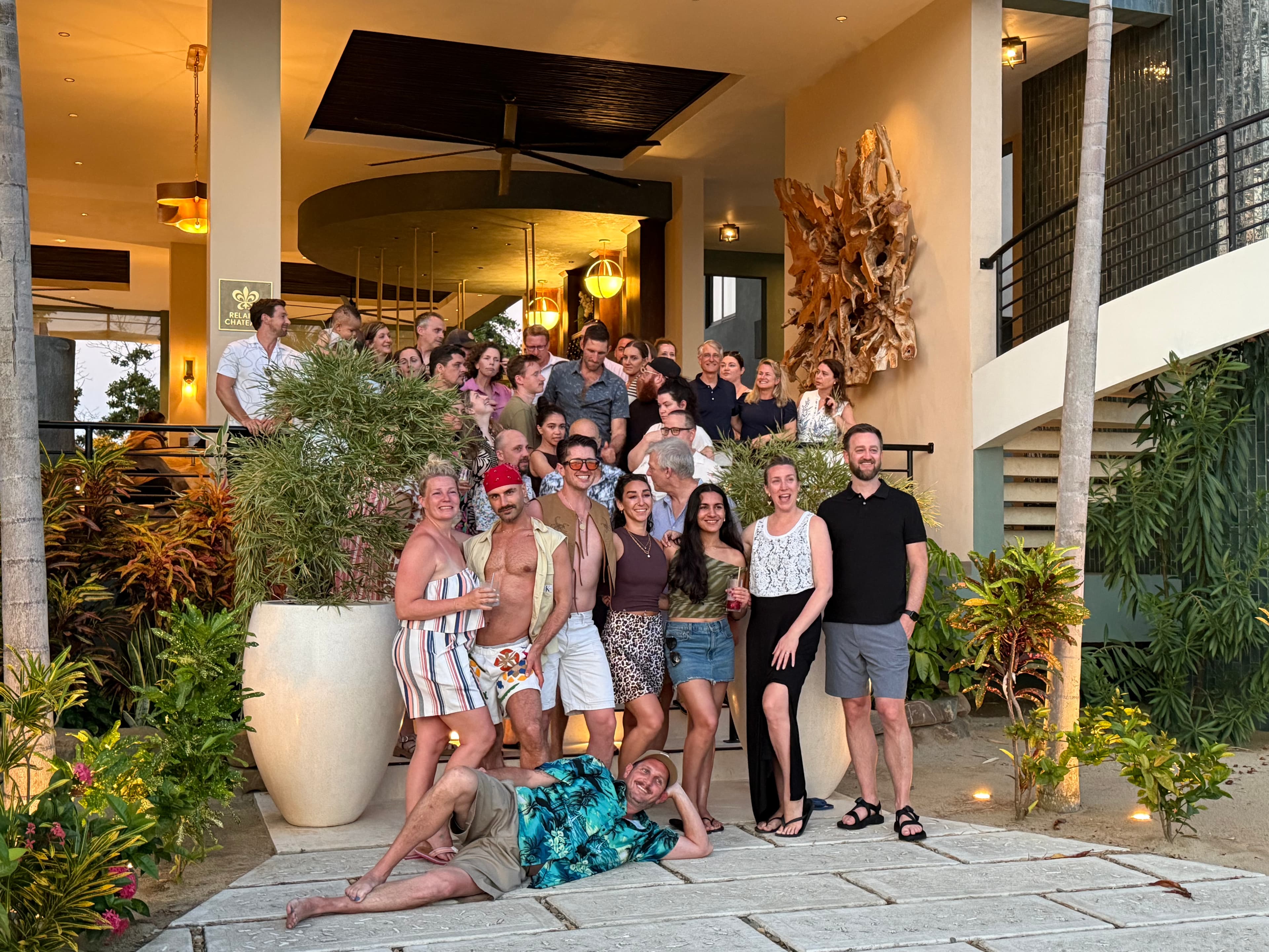 A large group of people posing together on a stone pathway in front of a modern building, surrounded by tropical plants.