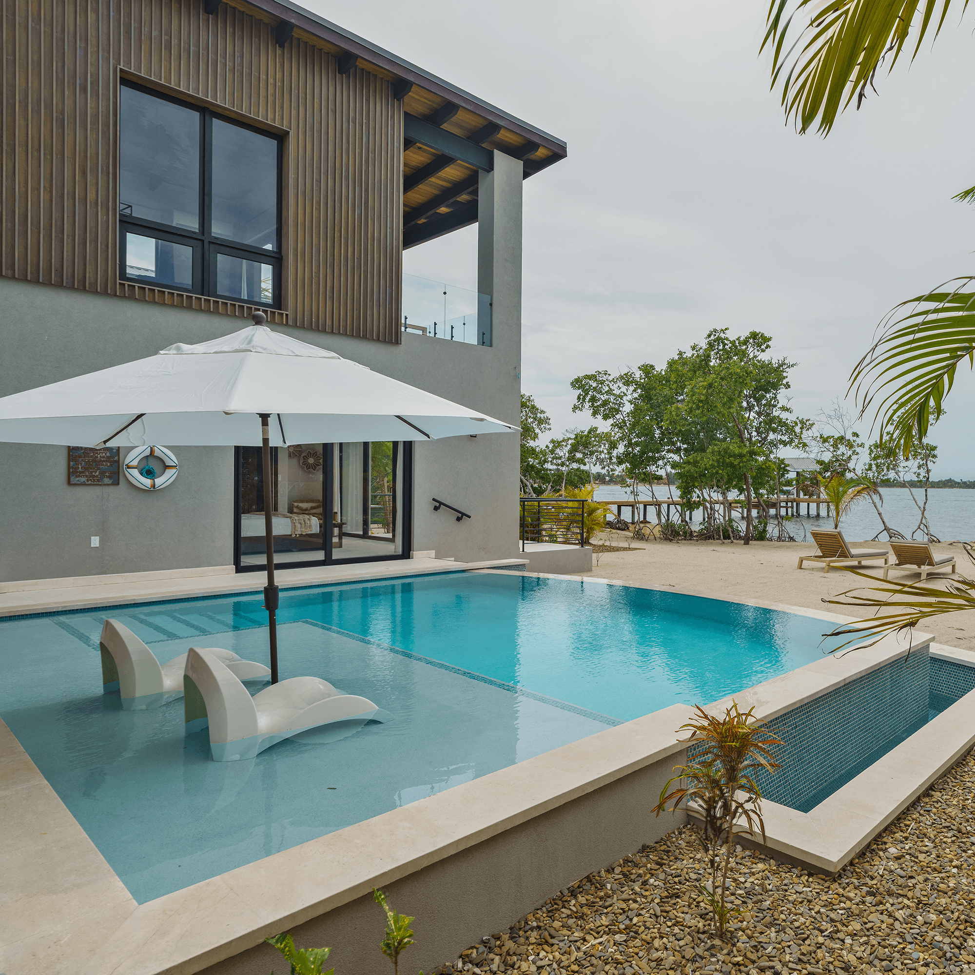A modern home featuring a swimming pool with lounge chairs and an umbrella, surrounded by tropical greenery and views of a lake.