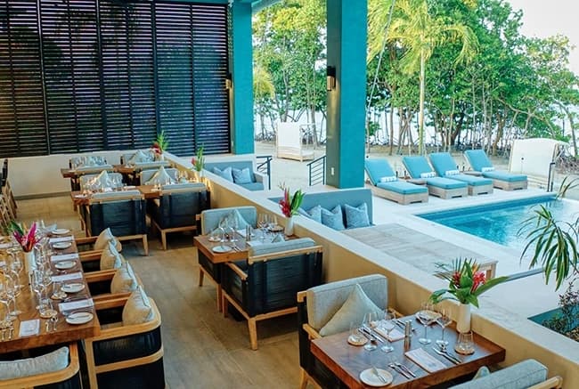 A stylish dining area with wooden tables and chairs overlooks a pool and lounge chairs surrounded by greenery.