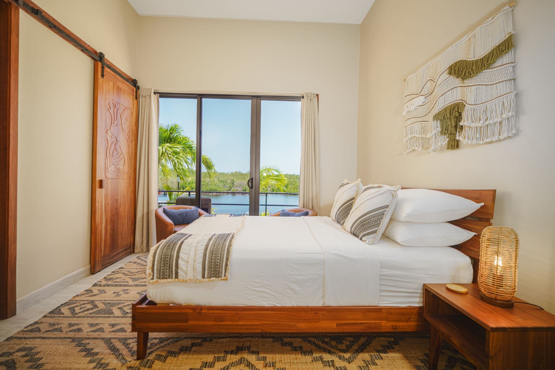 A serene bedroom featuring a wooden bed, decorative pillows, a woven wall hanging, and large windows with a view of greenery.