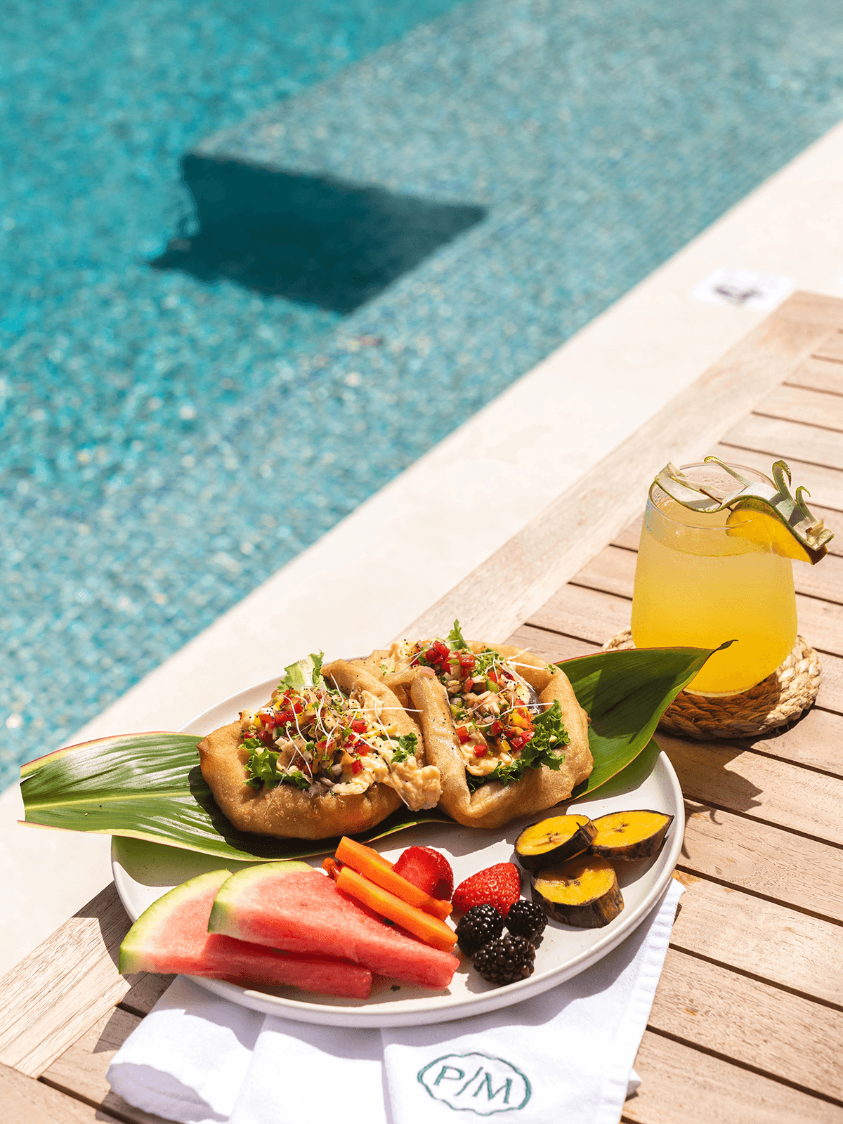 A plate of colorful food, including two filled tortillas, fresh fruits, and a drink, set beside a sparkling pool.