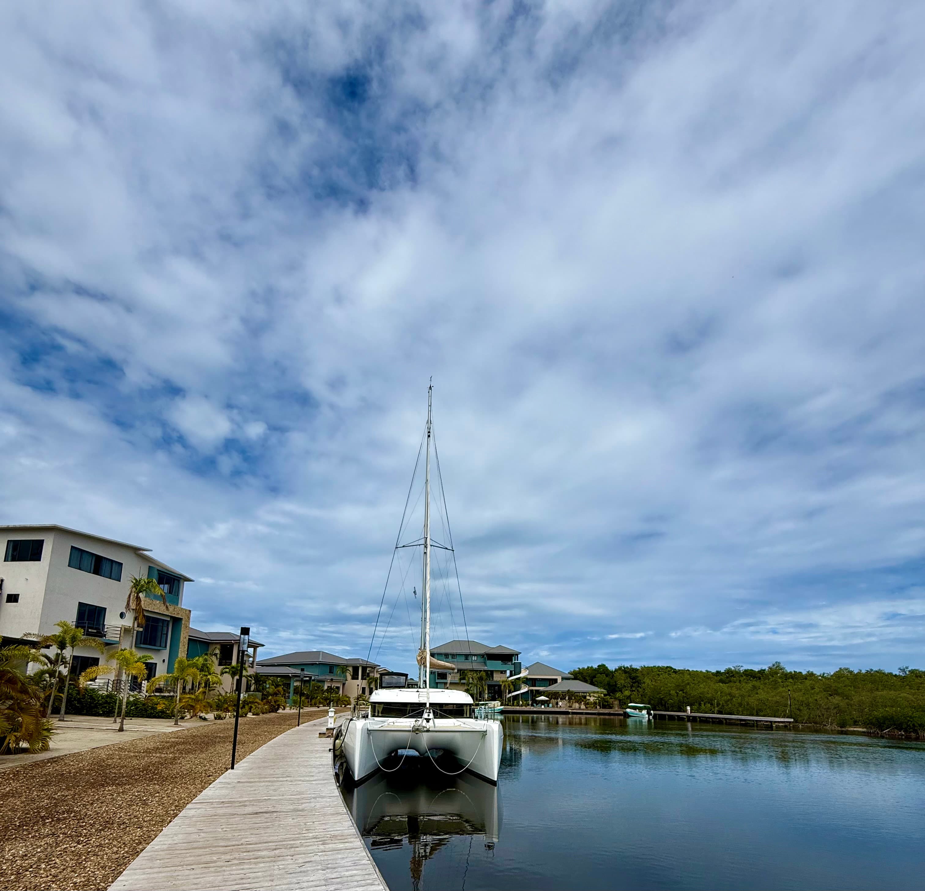 A catamaran is docked beside a wooden walkway with houses and distant greenery under a cloudy sky.