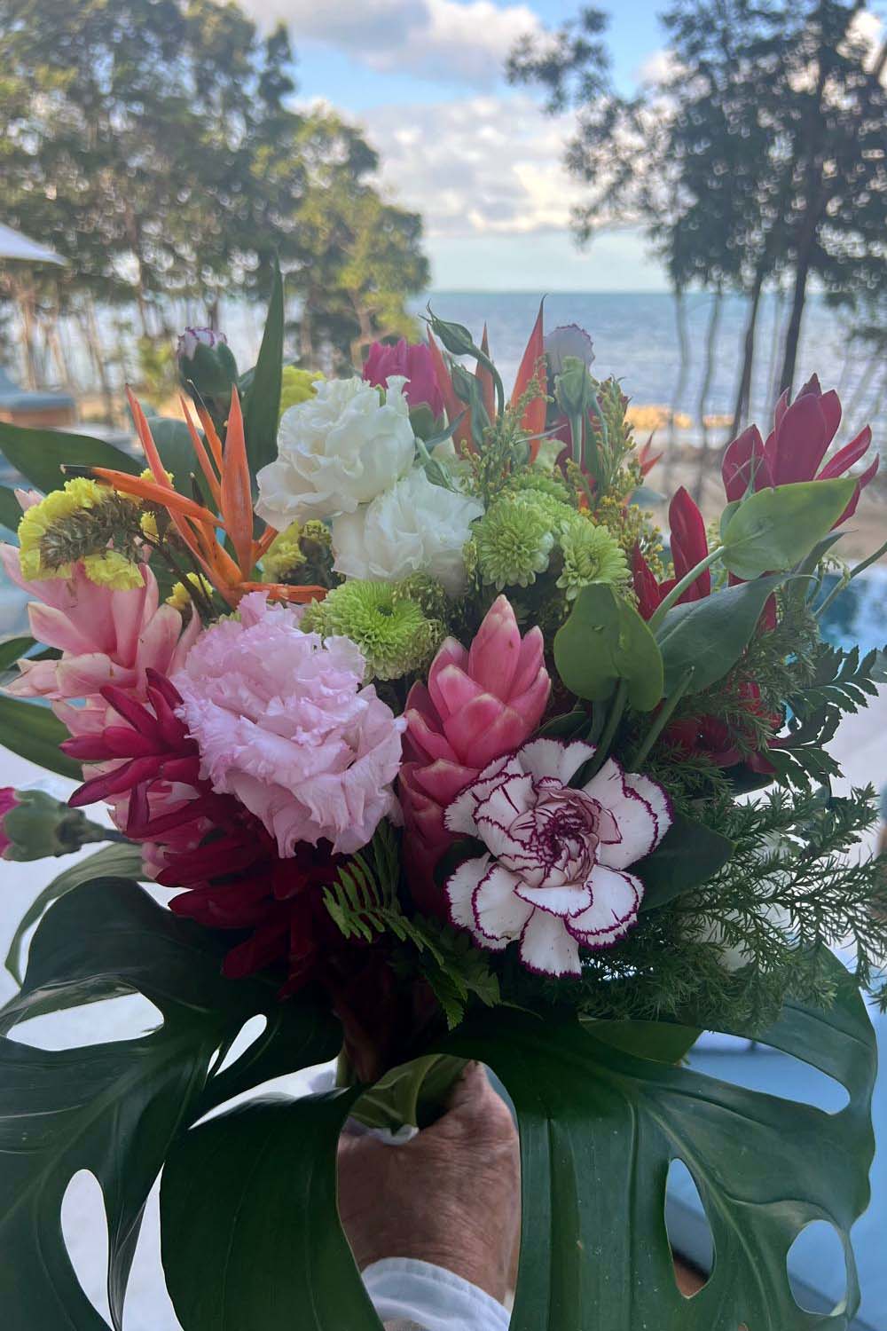 A hand holding a vibrant bouquet of mixed tropical flowers with a beach in the background.