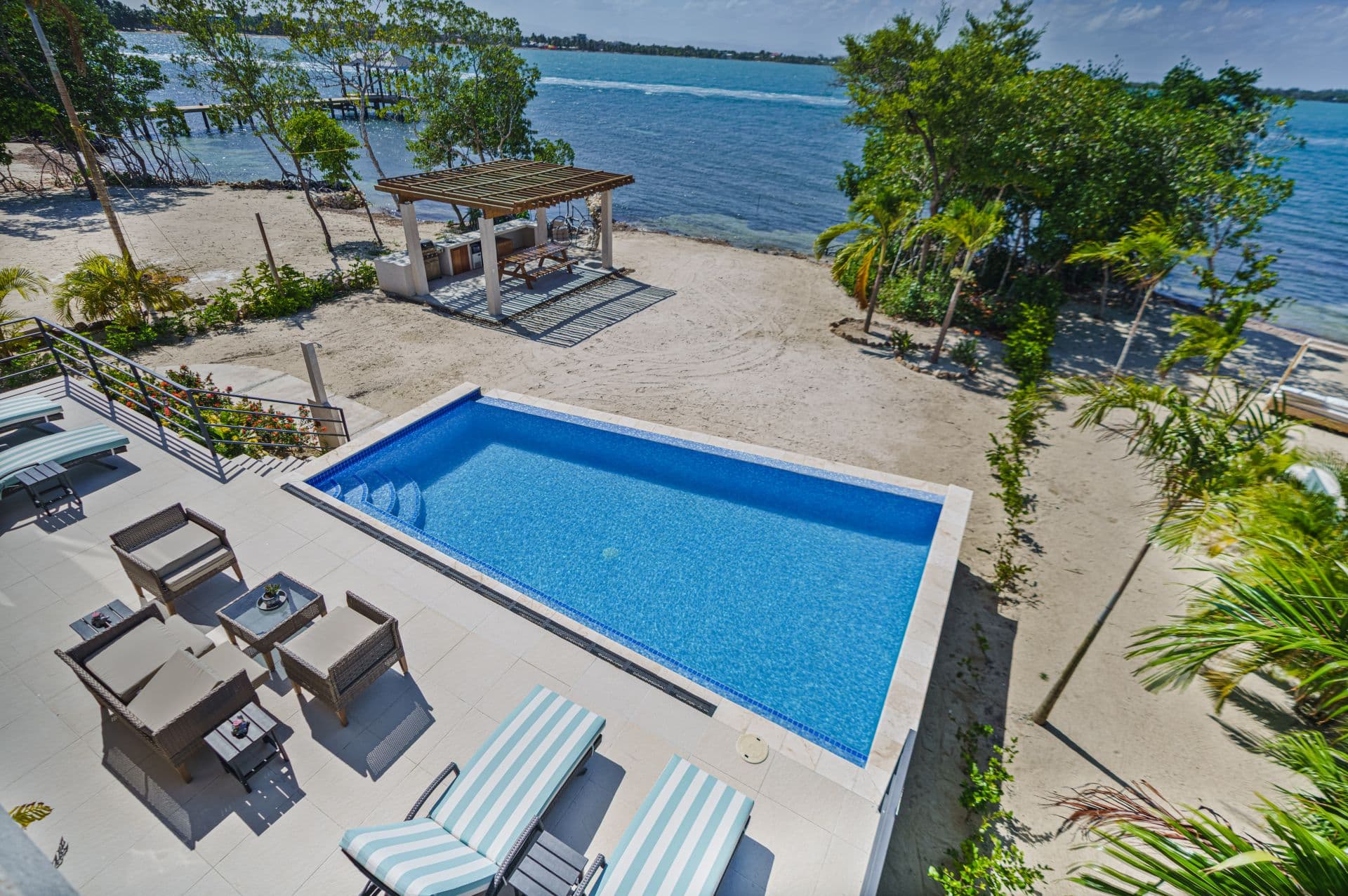 A panoramic view of a private pool overlooking a sandy beach and calm waters surrounded by lush greenery.