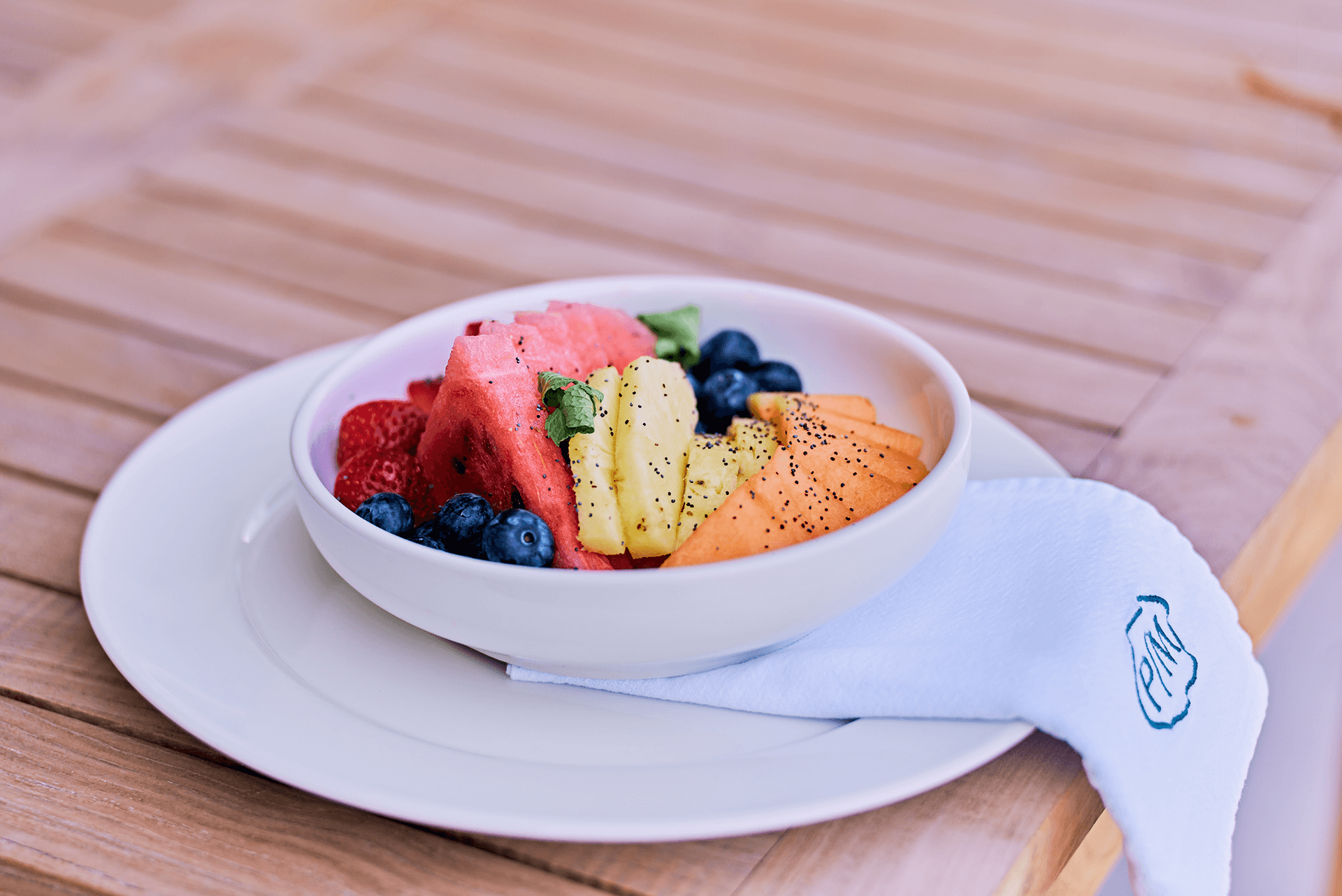 A bowl of mixed fresh fruit including watermelon, strawberries, pineapple, and blueberries on a wooden table.