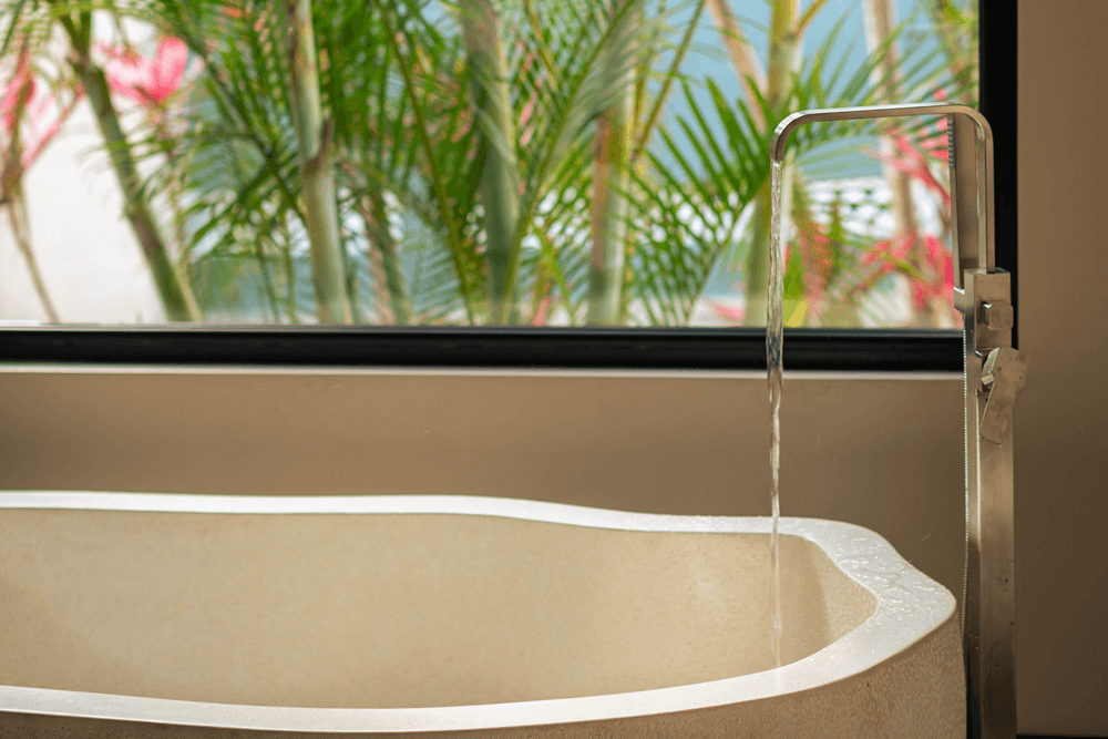 A modern bathtub with water flowing, set against a backdrop of plants visible through a window.