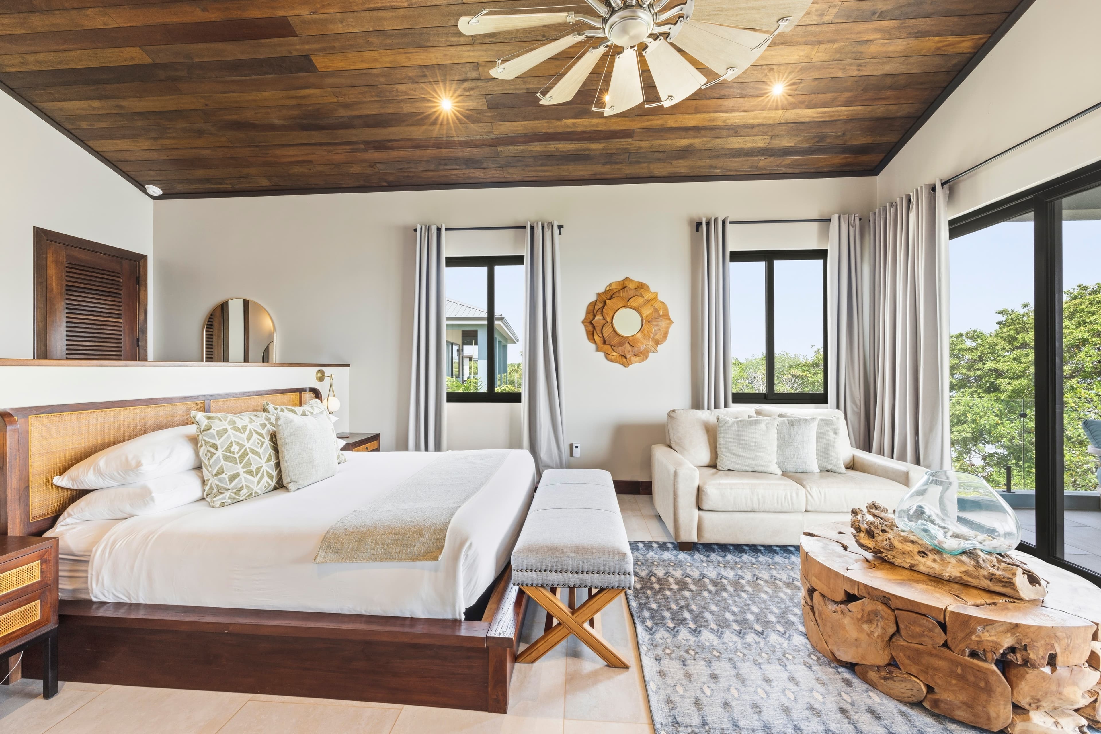 A stylish bedroom featuring a wooden ceiling, king-sized bed, cozy seating area, and large windows with a view.
