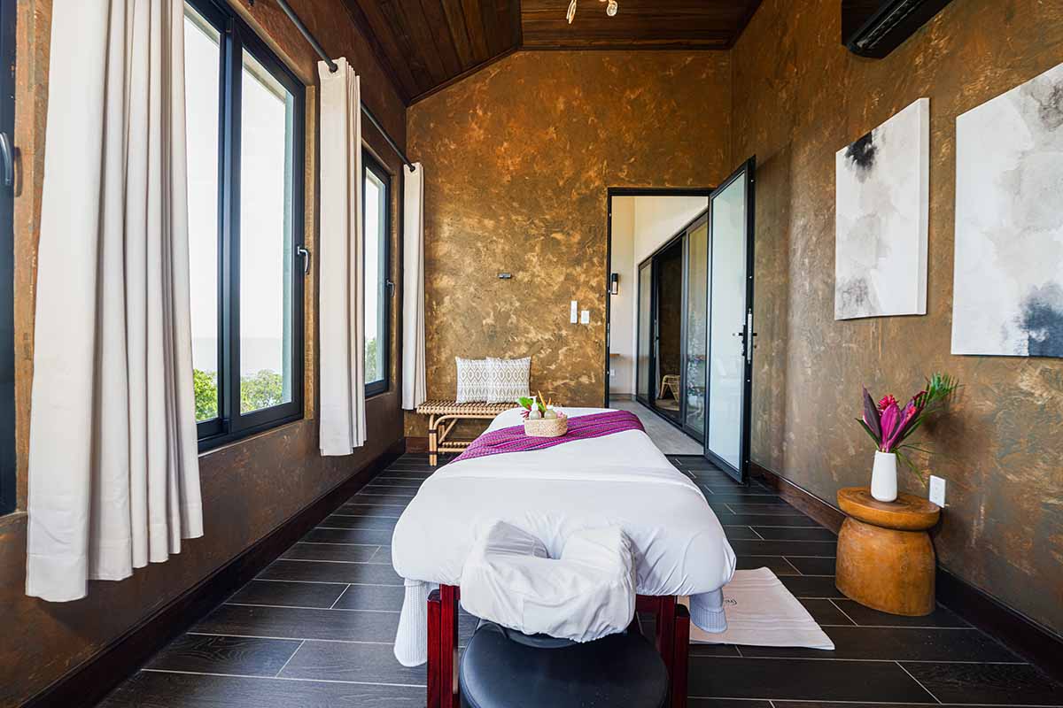 A serene massage room with large windows, a treatment table, and warm decor.