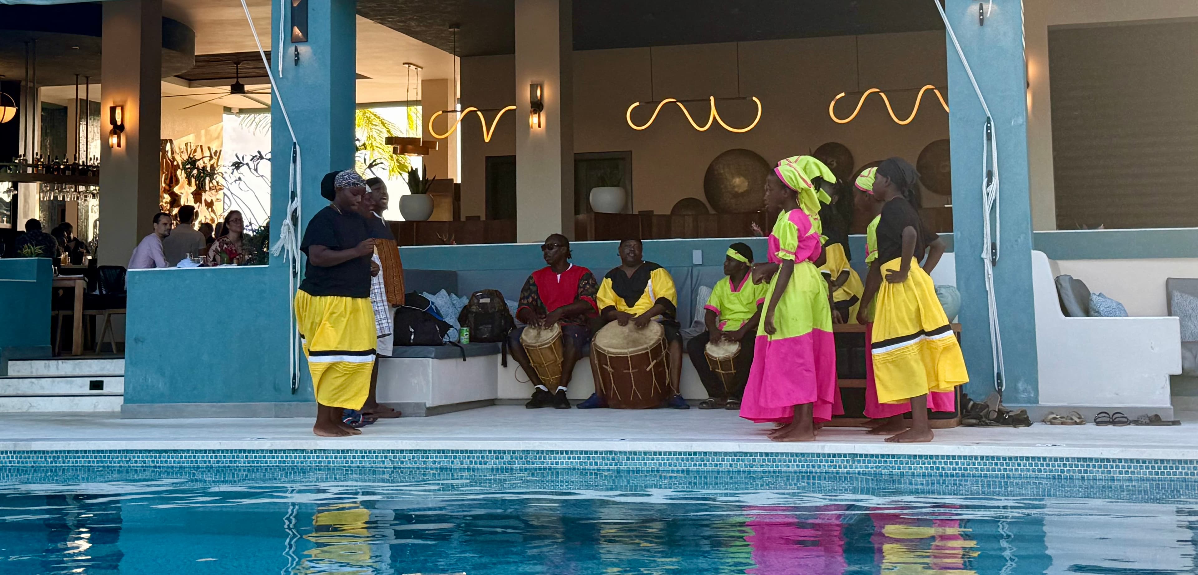A group of performers in vibrant costumes dance by a pool while drummers play in the background.