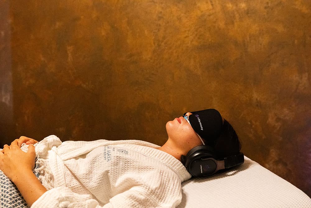 Person wearing headphones and a sleep mask lying on a bed against a gold-colored wall.