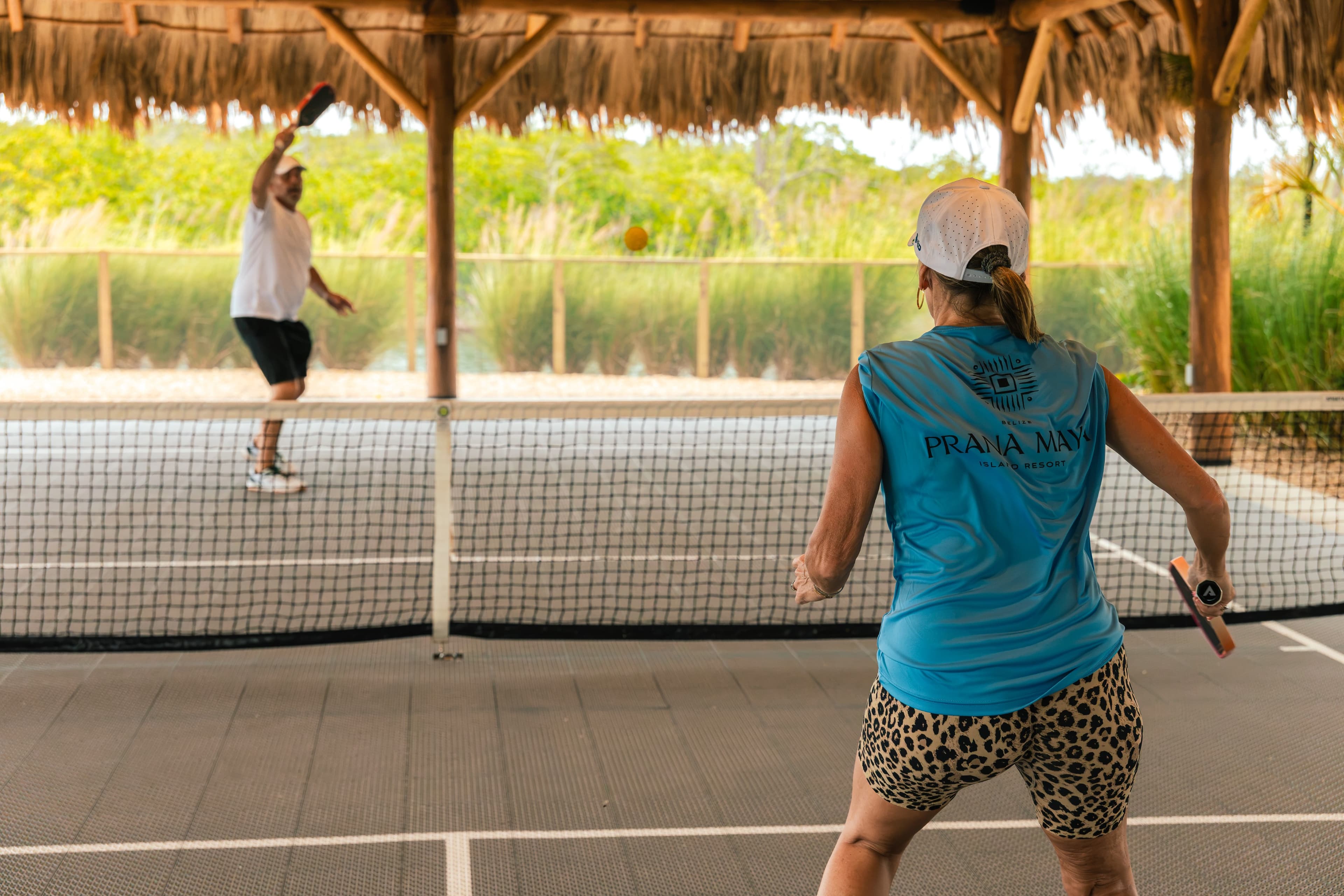 A woman in a blue tank top and leopard-print shorts prepares to return a ball while a man serves from the opposite side of a pickleball court.