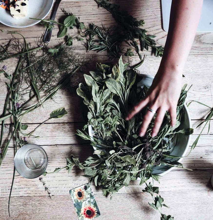 A hand reaches into a bowl filled with fresh herbs on a wooden table scattered with more herbs and a glass of water.