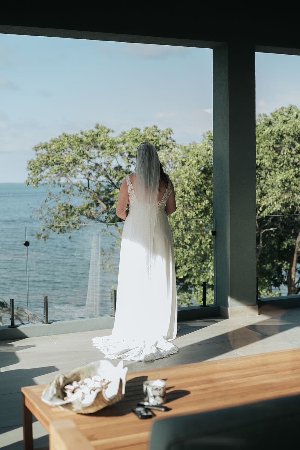A bride in a white gown stands with her back to the camera, overlooking a serene ocean view from a modern indoor space.