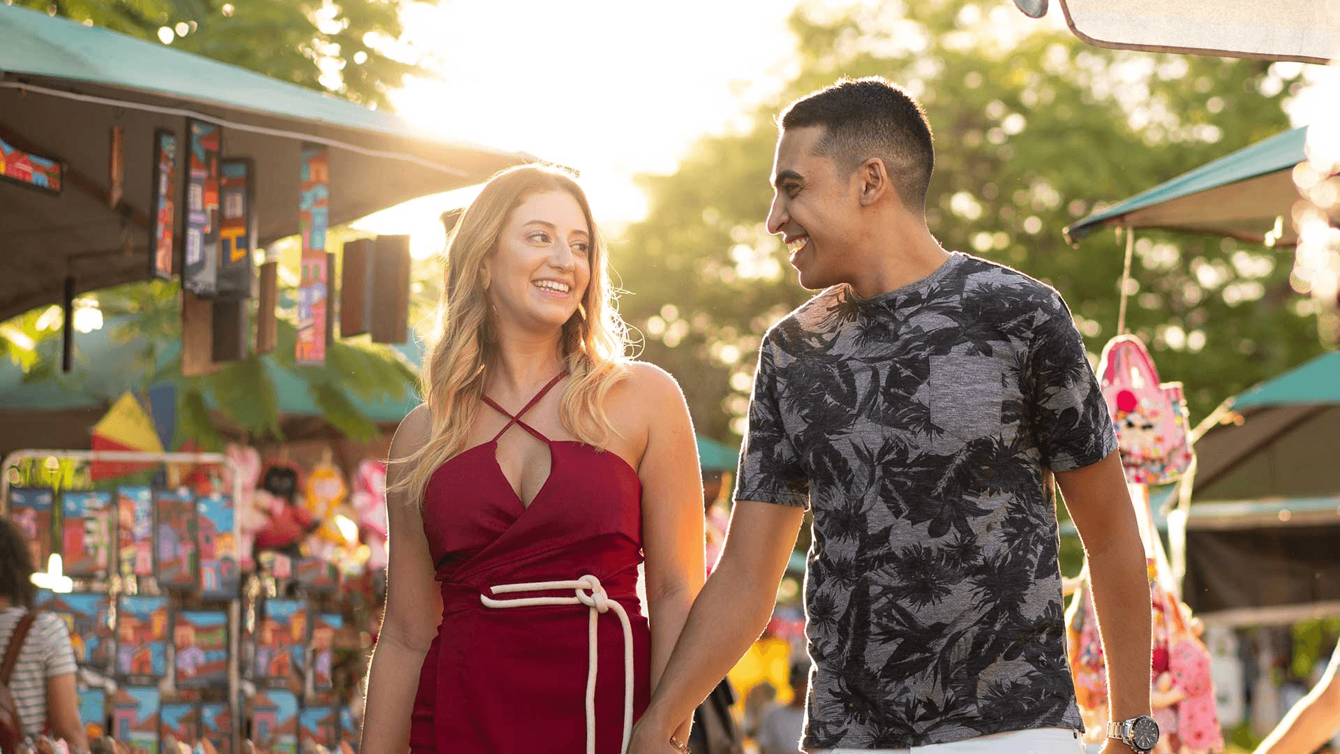 A young couple holds hands and smiles at each other while walking through a vibrant outdoor market at sunset.