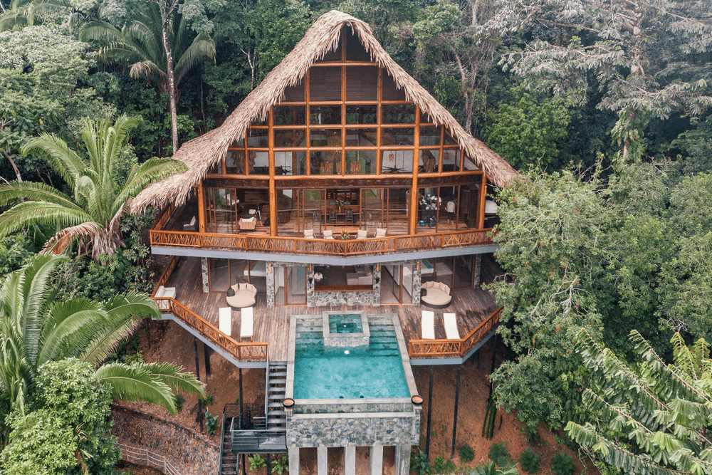 A modern, multi-level home with a thatched roof nestled in lush greenery, featuring a small pool on the terrace.