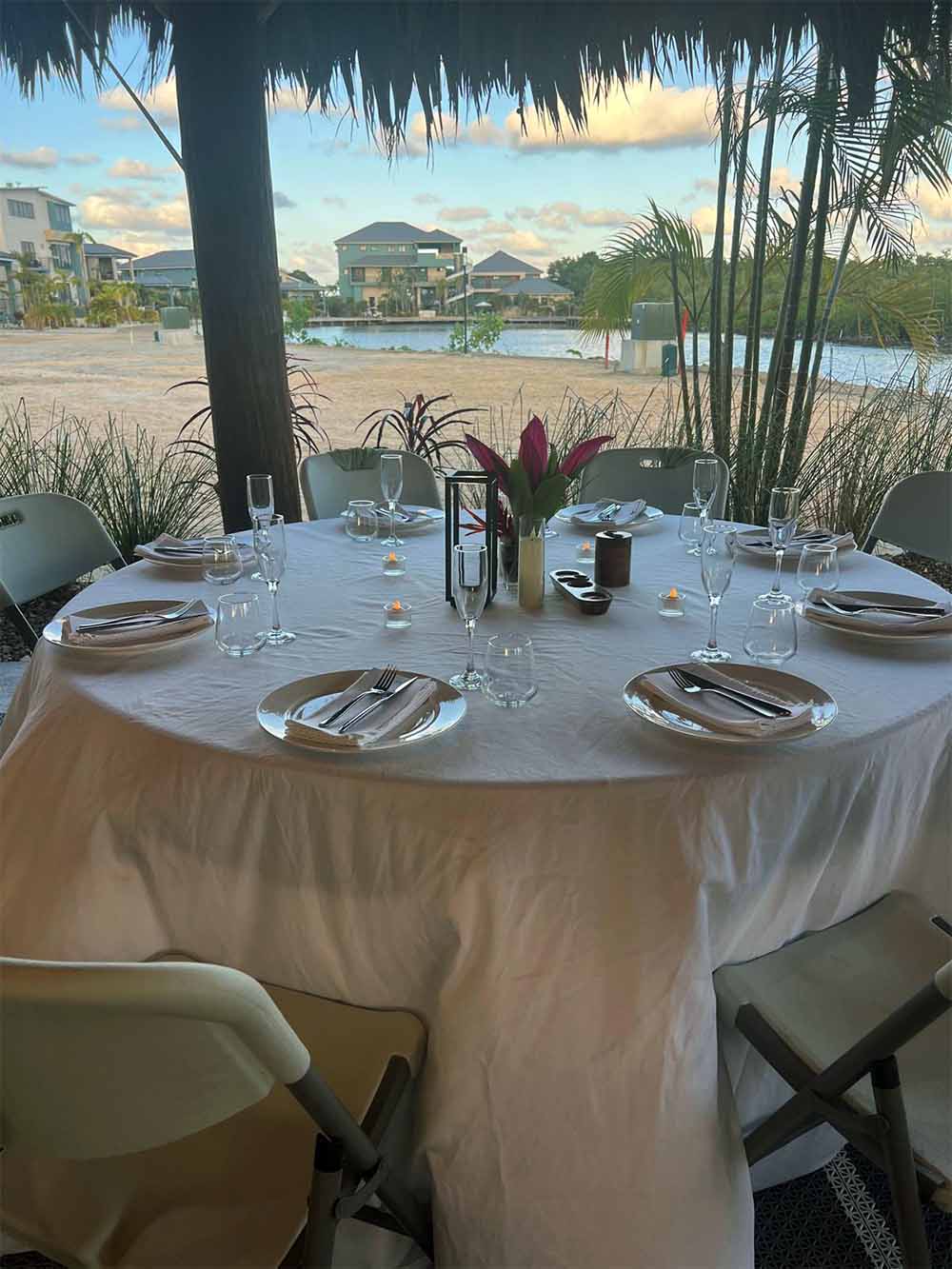 A beautifully set dining table with a view of a serene waterfront and tropical plants.