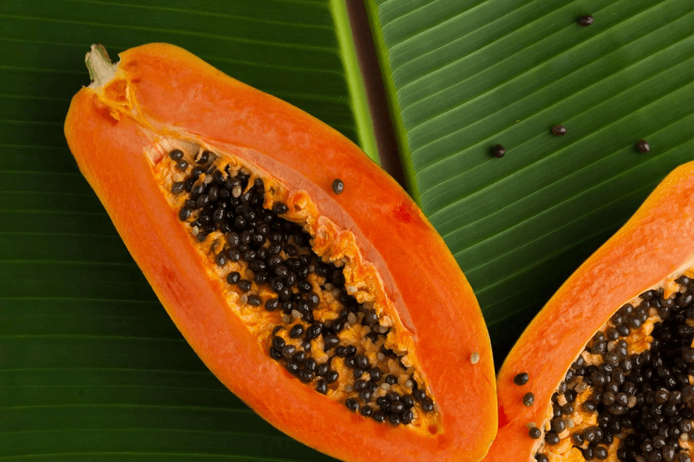Cut papaya displaying vibrant orange flesh and black seeds on green leaves.