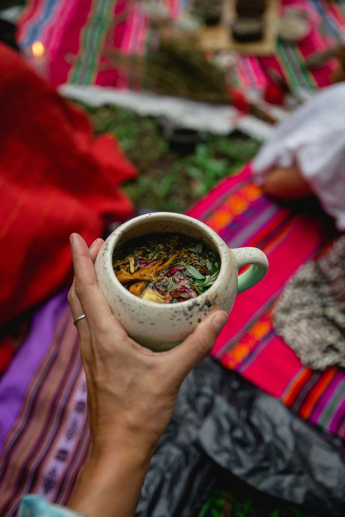 A hand holds a rustic, speckled mug filled with herbal tea. Colorful patterned blankets, in reds and pinks, cover the grass, creating a cozy picnic setting.