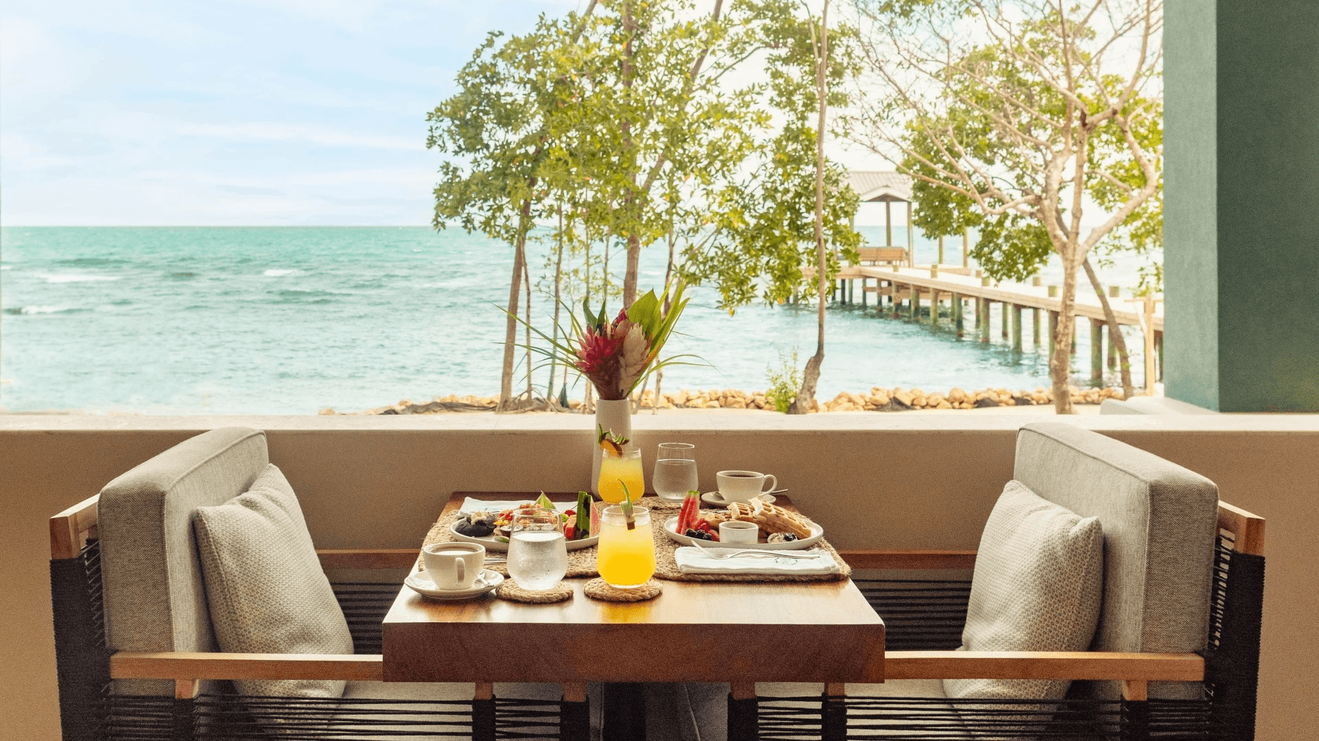 A serene seaside dining setting with two cushioned chairs, a wooden table with breakfast, vibrant flowers, and a view of the ocean and trees.
