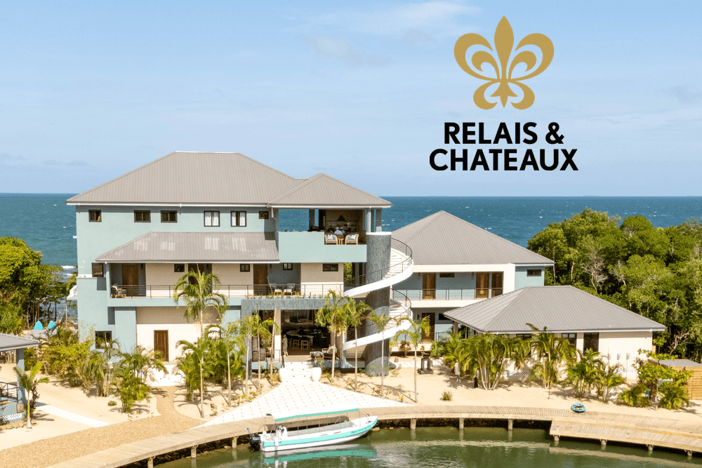 Oceanfront resort with pastel buildings, lush greenery, and a small dock with a boat. The mood is tranquil. "Relais & Chateaux" logo is visible.