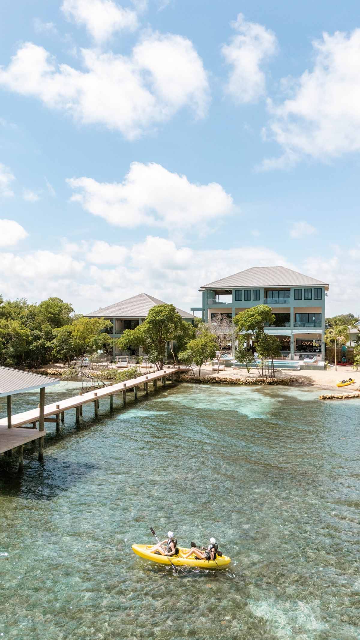 Two people kayaking on clear blue water near a pier, with modern buildings and lush trees in the background under a bright, partly cloudy sky.