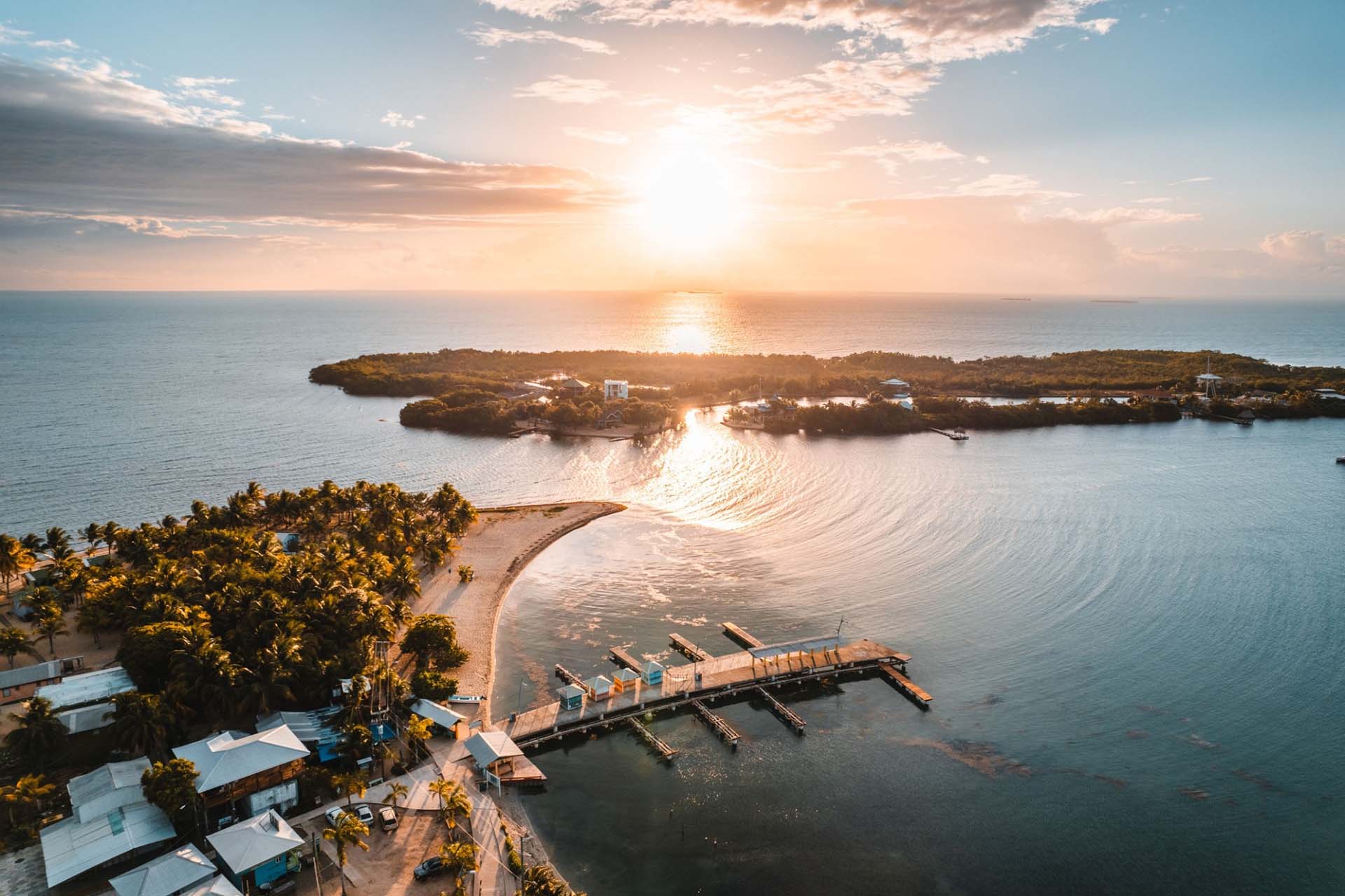 Aerial view of a serene coastal landscape at sunset. The sun casts a golden glow over the ocean, boats docked at a pier, and lush greenery nearby. Calm and tranquil.