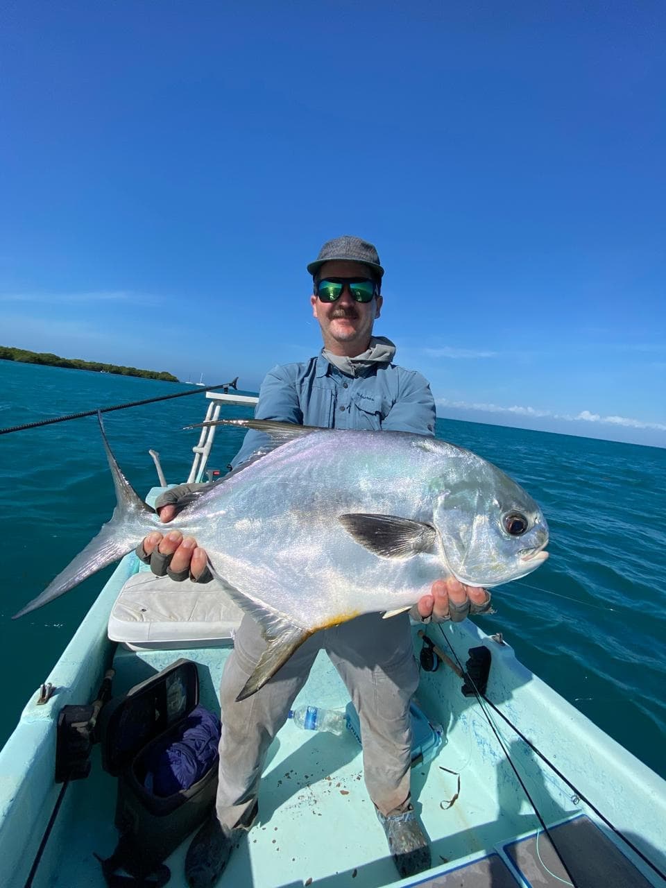 A person in sunglasses and a hat is smiling, holding a large, shiny fish on a boat in clear blue waters under a vivid blue sky.
