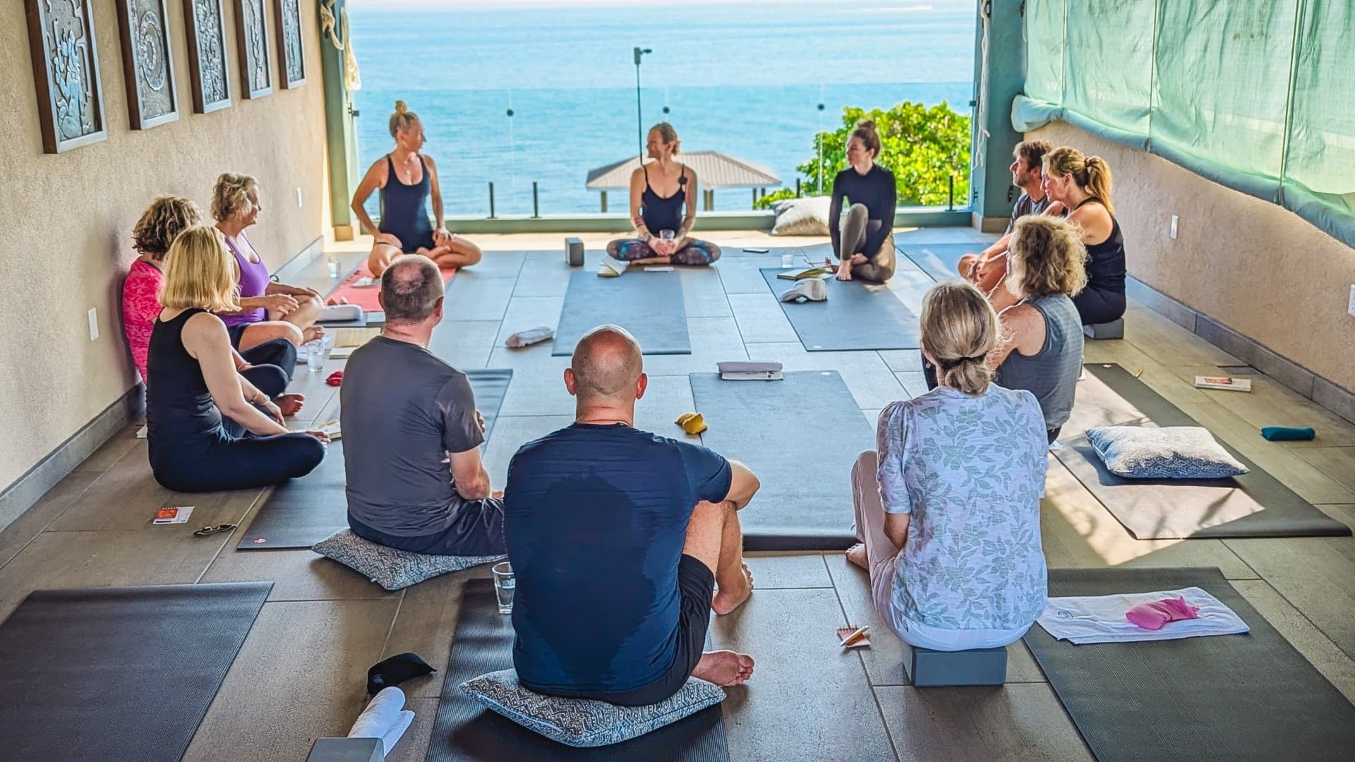 A serene yoga class with diverse participants seated on mats in a sunlit room overlooking the ocean. The instructor leads, fostering a calm and focused atmosphere.