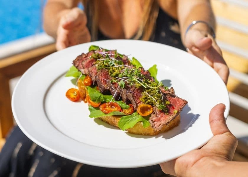 Guest holding a plated steak sandwich with fresh greens and cherry tomatoes poolside at Prana Maya Island Resort.