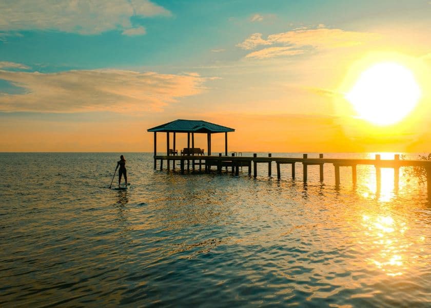 Silhouette of a person paddleboarding near a covered dock at Prana Maya Island Resort, at sunset over calm water. Silhouette of a person paddleboarding near a covered dock at Prana Maya Island Resort, at sunset over calm water.